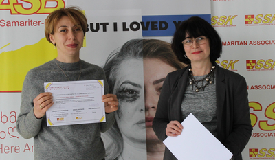 Zwei Frauen halten Zertifikate vor einem Banner mit einer emotionalen Botschaft.