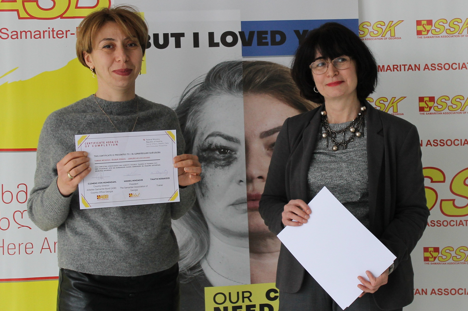 Zwei Frauen halten Zertifikate vor einem Banner mit einer emotionalen Botschaft.