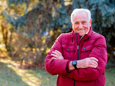 Ein lächelnder älterer Mann in einer roten Jacke steht mit verschränkten Armen.