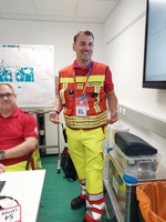 Ein Rettungskräfte-Uniformierter steht im Vordergrund und lächelt, während er spricht. Im Hintergrund sitzt eine weitere Person. Beide tragen auffällige, reflektierende Kleidung in Rot und Gelb. An einer Wand ist eine Karte sichtbar. Diverse Ausrüstungsgegenstände sind im Raum verteilt.