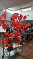 Rote Herzluftballons in einem Raum mit älteren Menschen, die aufmerksam zuhören.