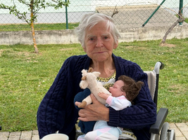 Eine ältere Frau im Rollstuhl sitzt an einem Tisch im Freien. Sie hält eine Puppe und ein Stofftier in den Händen. Auf dem Tisch vor ihr stehen eine Tasse und ein Stück Kuchen. Im Hintergrund ist eine grüne Wiese zu sehen.