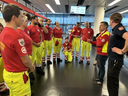 Eine Gruppe von Rettungskräften in rot-gelben Uniformen versammelt sich in einem modernen Raum. Ein Teamleiter spricht. Einige Teammitglieder hören aufmerksam zu, während andere eine entspannte Haltung einnehmen. Im Hintergrund sind große Fenster mit Stadtansicht sichtbar.