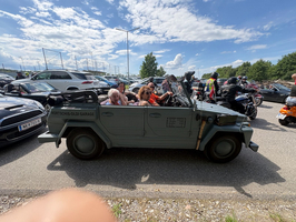 Eine Gruppe von Menschen sitzt in einem alten, grünen Auto im offenen Design. Das Fahrzeug ist umgeben von moderneren Autos und Motorrädern auf einem Parkplatz, unter einem klaren, blauen Himmel mit vereinzelten Wolken.