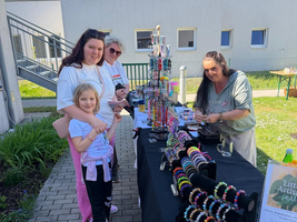Drei Personen stehen an einem Tisch mit handgemachtem Schmuck, während eine Frau am Tisch arbeitet. Auf dem Tisch sind unterschiedliche Armbänder und andere Schmuckstücke ausgestellt. Im Hintergrund sind grüne Wiesen und ein Gebäude sichtbar. Es ist ein sonniger Tag.