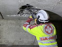 Ein Arbeiter in Schutzkleidung mit einem Helmut und einer Warnjacke bearbeitet eine beschädigte Betondecke. Er benutzt ein Werkzeug, um eine Öffnung im Beton zu erweitern, während er auf den nächsten Schritt vorbereitet ist.
