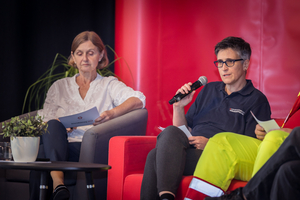 Zwei Frauen auf einem Podium, eine spricht ins Mikrofon, die andere hört zu.