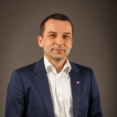 Ein Mann in einem blauen Anzug mit offenem Hemd steht vor einem neutralen Hintergrund.