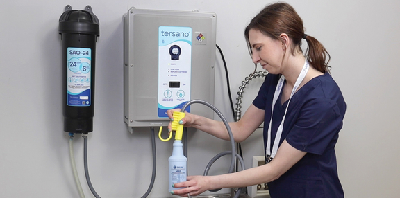 Eine Person in blauer Arbeitskleidung hält eine Sprühflasche und befüllt sie an einem Waschbecken. An der Wand ist ein Gerät zur Wasserbehandlung montiert, das mit Schläuchen verbunden ist. Im Hintergrund sind weitere Geräte sichtbar. Die Reinigung erfolgt ohne Chemie.