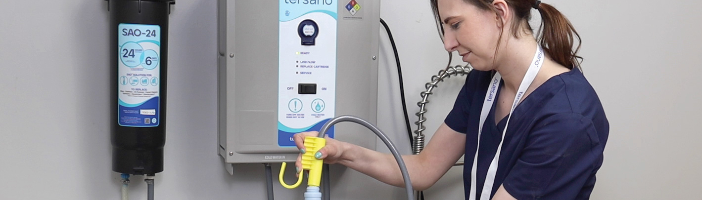 Eine Person in blauer Arbeitskleidung hält eine Sprühflasche und befüllt sie an einem Waschbecken. An der Wand ist ein Gerät zur Wasserbehandlung montiert, das mit Schläuchen verbunden ist. Im Hintergrund sind weitere Geräte sichtbar. Die Reinigung erfolgt ohne Chemie.