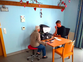 Eine ältere Frau sitzt an einem Tisch und nutzt ein Hilfsmittel mit einem Mann.