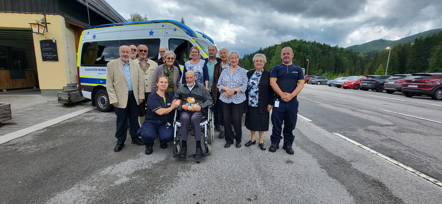 Eine Gruppe von zwölf Personen steht in einem Parkplatz vor einem Krankenwagen. Ein älterer Mann sitzt im Rollstuhl. Die Mitglieder der Gruppe sind unterschiedlich gekleidet, und die Umgebung zeigt eine bewaldete Landschaft unter einem bewölkten Himmel.