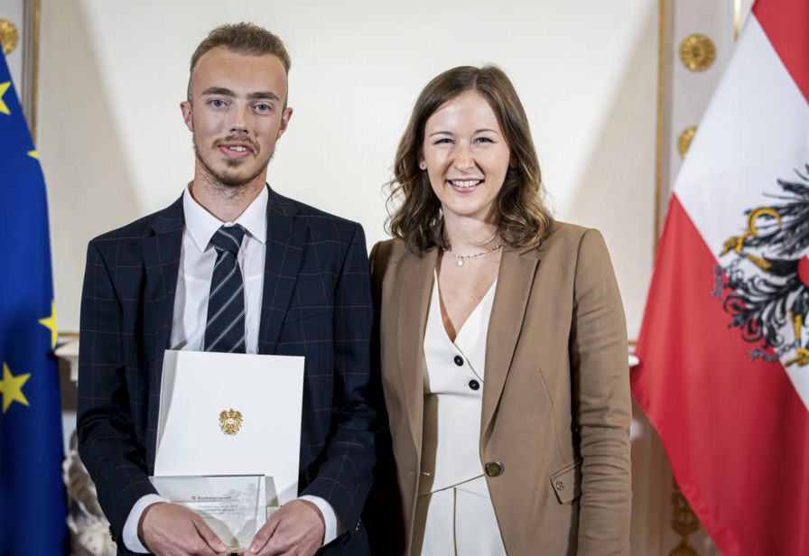 Zwei Personen posieren mit einer Urkunde vor den Flaggen der EU und Österreich.