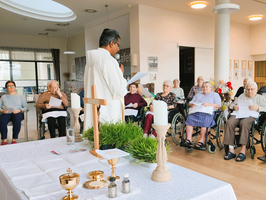 Ein Priester steht an einem Tisch mit religiösen Utensilien und spricht zu einer Gruppe von Menschen in Rollstühlen. Die Anwesenden halten gesammelte Lieder oder Texte in den Händen. Im Hintergrund sind dekorative Elemente und Fenster sichtbar.