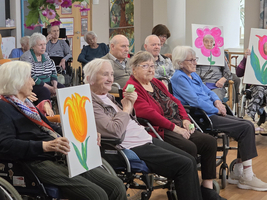 Eine Gruppe von älteren Menschen in Rollstühlen sitzt im Raum und hält gemalte Bilder von Blumen in den Händen. Einige Personen lachen oder lächeln, während sie an einem sozialen oder kreativen Aktivitätsprogramm teilnehmen. Im Hintergrund sind weitere Teilnehmer und Bilder an der Wand zu sehen.
