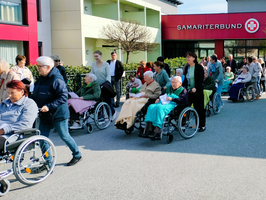 Eine Vielzahl von älteren Menschen in Rollstühlen steht in einer Schlange vor einem Gebäude mit dem Schild 