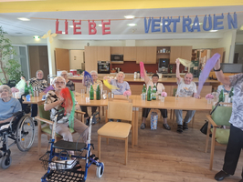 Eine Gruppe älterer Menschen sitzt an einem Tisch in einem Raum. Einige halten bunte Tücher hoch, während sie fröhlich lächeln. Im Hintergrund hängen bunte Banner mit den Worten 