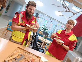 Zwei Rettungskräfte in roten und gelben Uniformen stehen in einem Raum. Sie halten Teller mit Essen in den Händen. Auf einem Tisch vor ihnen liegt ein halbierter Pizzastück und eine Küchenausrüstung. Der Raum ist hell und freundlich gestaltet, mit Pflanzen und bunten Zeichnungen im Hintergrund.