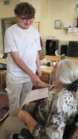 Ein junger Mann in einem weißen Oberteil spricht freundlich mit einer älteren Frau, die in einem Rollstuhl sitzt. Sie hält ein Blatt Papier in der Hand. Der Hintergrund zeigt einen Raum mit Tisch und verschiedenen Gegenständen.