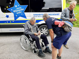 Ein älterer Mann im Rollstuhl erhält Hilfe von zwei Personen.
