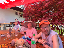 Eine Gruppe von vier Personen sitzt an einem Holztisch unter einem roten Sonnenschirm. Eine ältere Frau mit einem Hut lächelt in die Kamera, während die anderen in entspannter Pose neben ihr sitzen. Auf dem Tisch liegen einige Spielzeuge und eine Flasche Wasser.