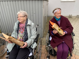 Zwei ältere Frauen in Rollstühlen halten Holzstücke mit dekorativen Elementen.