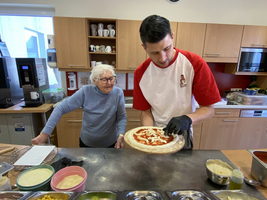 Eine ältere Frau schaut zu, während ein Mann Pizza belegt.