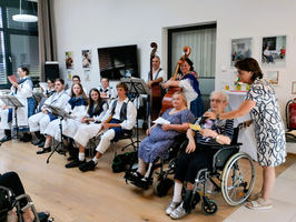 Eine Gruppe von Musikern in traditionellen Kostümen spielt auf einer Bühne, während einige ältere Menschen im Publikum sitzen. Eine Frau mit einem Getränk spricht mit einer der Damen im Rollstuhl. Im Hintergrund sind Fotos an der Wand zu sehen.
