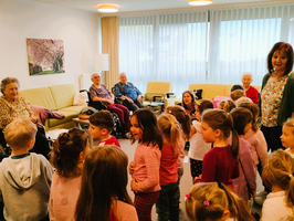 Eine Gruppe von Kindern steht in einem Wohnraum und singt oder spielt für mehrere ältere Menschen, die auf Sofas sitzen. Einige Senioren lächeln, während die Kinder mit Freude interagieren. Der Raum ist hell und freundlich, mit Fenster und Bildern an den Wänden.