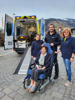 Vier Personen stehen neben einem Krankenwagen; eine ältere Frau sitzt auf einem Fahrstuhl.