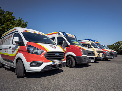 Fünf Rettungsfahrzeuge stehen auf einem Parkplatz in einer Reihe, darunter ein rotes, weißes und gelbes Fahrzeug. Die Fahrzeuge sind in verschiedenen Farben gestaltet und tragen aufgedruckte Logos. Im Hintergrund sind Bäume und ein blauer Himmel sichtbar.