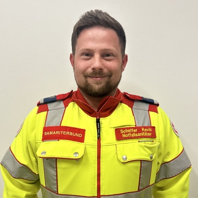 Mann in gelber Sanitäteruniform mit lächelndem Gesicht vor neutralem Hintergrund.