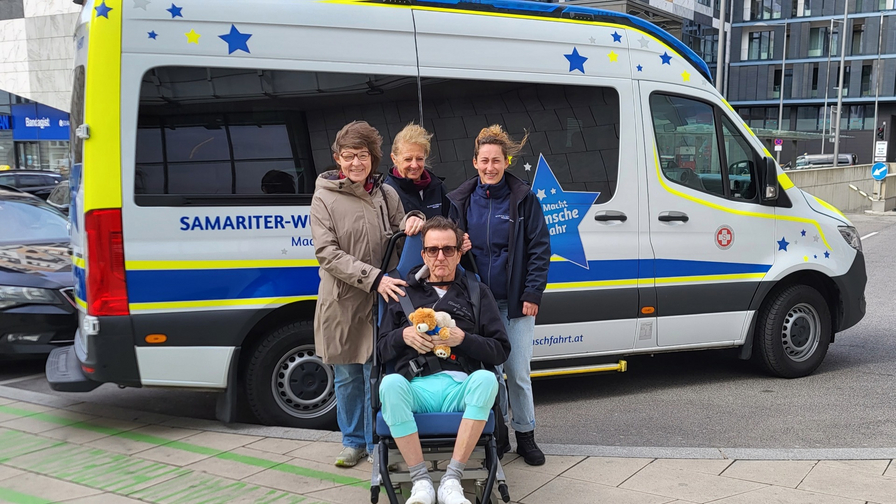 Vier Personen stehen vor einem gelben und blauen Fahrzeug mit dem Aufdruck „Samariter-Wagen“. Eine Person sitzt in einem Rollstuhl und hält ein Kuscheltier. Alle Personen lächeln in die Kamera, während sie eine freundliche Atmosphäre ausstrahlen.