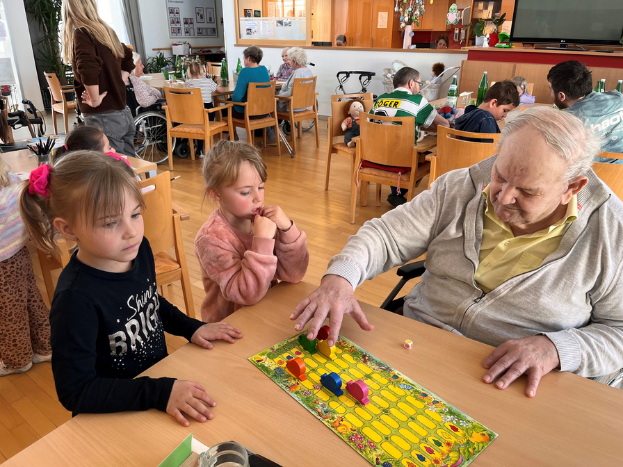 Auf dem Bild sind zwei Mädchen und ein älterer Mann zu sehen, die an einem Tisch spielen. Der Mann bewegt Spielfiguren auf einem bunten Spielbrett. Im Hintergrund sind weitere Personen in einem Gemeinschaftsraum sichtbar, die ebenfalls beschäftigt sind.
