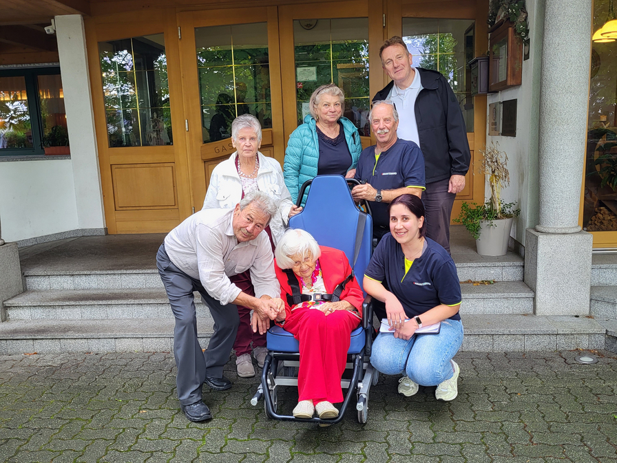 Eine Gruppe von sieben Personen posiert vor einem Gebäude mit einer Seniorin im Rollstuhl.