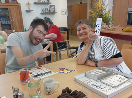 Zwei Personen sitzen an einem Tisch in einem Gemeinschaftsraum. Eine Frau hält eine Bingo-Karte mit der Zahl G47 hoch, während ein Mann lächelt und mit dem Finger auf sie zeigt. Im Hintergrund sind weitere Personen und verschiedene Spielsachen zu sehen. Getränke und Snacks liegen auf dem Tisch.