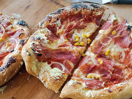Zwei frisch gebackene Pizzen auf einem Holzbrett, eine in Stücke geschnitten. Die Pizzen sind mit Tomatensauce, Käse, Schinken, Zwiebeln und Mais belegt. Im Hintergrund sind Küchenutensilien sichtbar.