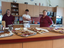 Drei Personen stehen in einer Küche mit verschiedenen Kuchen und Snacks auf dem Tisch.