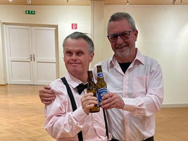 Zwei Männer in Hemden halten Bierflaschen und lächeln in freundlicher Atmosphäre.