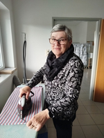 Eine ältere Frau mit Brille und einem schwarzen Schal bügelt ein gestreiftes Tuch an einem Bügeltisch. Im Hintergrund sind weitere Textilien und eine Tür zu sehen.
