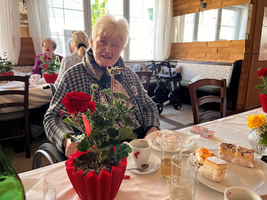 Eine ältere Frau sitzt an einem Tisch mit Blumen und Kuchen.