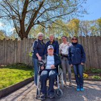 Eine Gruppe von fünf Personen steht im Freien vor einem Holzzaun. Eine Person sitzt in einem Rollstuhl, während die anderen um ihn herum stehen und lächeln. Im Hintergrund sind Bäume und grünes Gras zu sehen. Es ist sonnig und der Himmel ist blau.