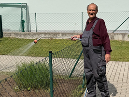 Ein älterer Mann steht am Zaun eines Gartens und spritzt Wasser mit einem Gartenschlauch. Er trägt eine lockere Kleidung und lächelt. Im Hintergrund sind Gebäude und eine grüne Wiese zu sehen.