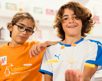 Zwei Kinder aus dem LernLEO blicken in Kamera und lächeln, Mädchen mit Brille und orangefarbenem T-Shirt lehnt an Jungen mit längeren Haaren und weiß,blauen T-Shirt der einen Obstspieß in die Kamera hält und grinst