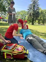 Eine Gruppe von Kindern übt Erste Hilfe im Freien. Ein Junge liegt auf einer Decke und wird von einem Mädchen behandelt, während ein Erwachsener und zwei weitere Kinder zuschauen. Im Vordergrund liegt eine rote Erste-Hilfe-Tasche.