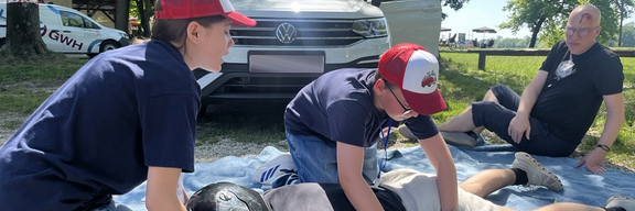 Zwei Kinder leisten erste Hilfe an einer liegenden Person auf einer Decke. Die Person trägt einen Helm und scheint verletzt zu sein. Im Hintergrund steht ein Auto, während eine weitere Person zuschaut. Bäume und grünes Gras sind sichtbar.