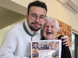 Zwei Personen stehen in einem Innenraum zusammen. Eine junge Person hält ein gerahmtes Plakat mit Bildern und Text in den Händen. Eine ältere Frau lächelt und umarmt den jungen Mann. Beide sind fröhlich und scheinen einen besonderen Moment zu teilen.