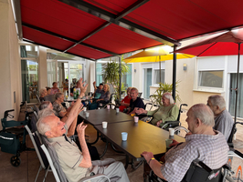 Eine Gruppe älterer Menschen sitzt an einem Tisch unter roten Sonnenschirmen. Einige Personen sprechen lebhaft und gestikulieren, während andere aufmerksam zuhören. Im Hintergrund sind Pflanzen und ein hell erleuchteter Raum sichtbar.