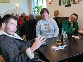 Eine Gruppe von vier Personen sitzt in einem Café. Ein junger Mann im Rollstuhl macht eine Geste mit seiner Hand. Neben ihm sitzt eine Frau mit einem grauen Sweatshirt. Im Hintergrund sind ältere Menschen zu sehen, die an Tischen sitzen.