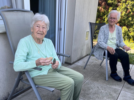 Zwei ältere Frauen sitzen auf einer Terrasse. Eine Frau trägt hellgrüne Kleidung und hält etwas in der Hand, während die andere in einem grauen Oberteil sitzt. Neben ihnen stehen Papiertragetaschen. Im Hintergrund sind Bäume und eine Wand zu sehen.